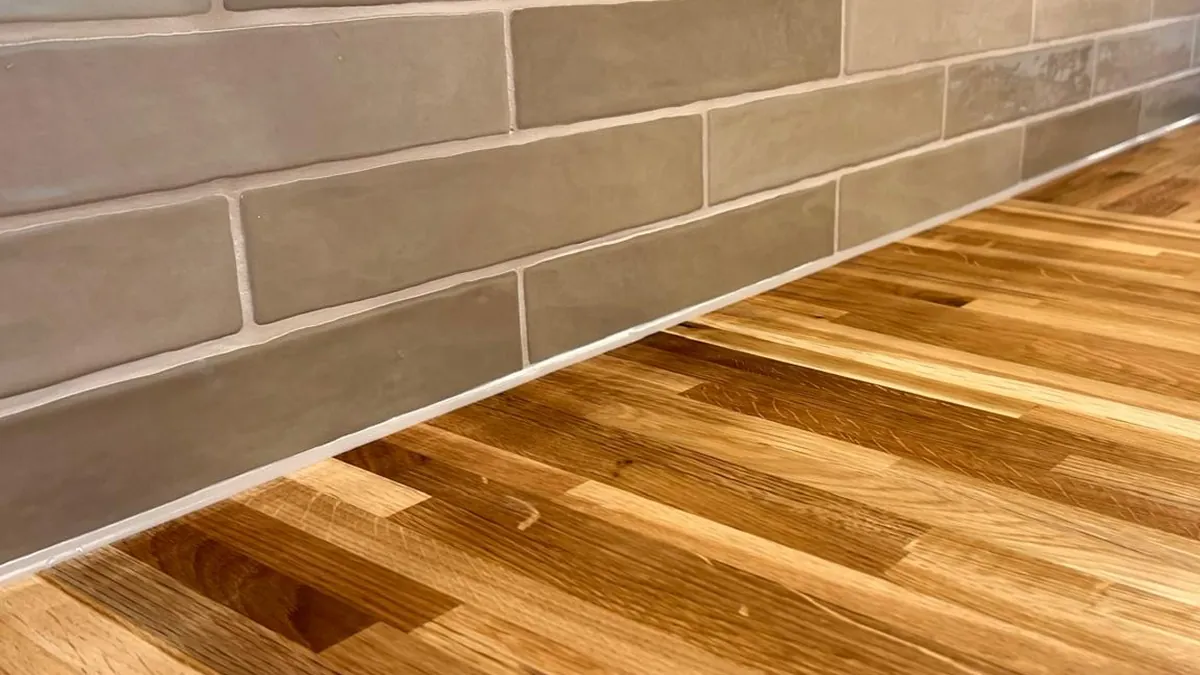 Thin pale silicone joint along the back of a solid oak kitchen worktop where it meets grey brick-shaped wall tiles