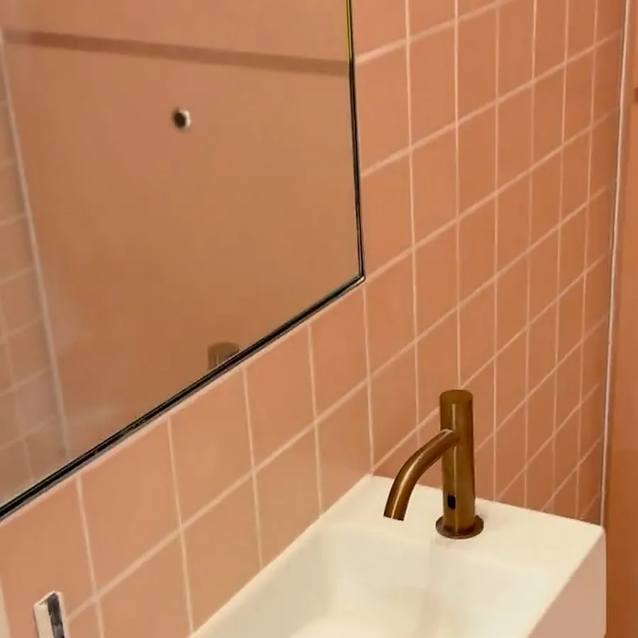 Pink square wall tiles, a frameless mirror and a bronze sensor tap above the back edge of a white wall-hung basin