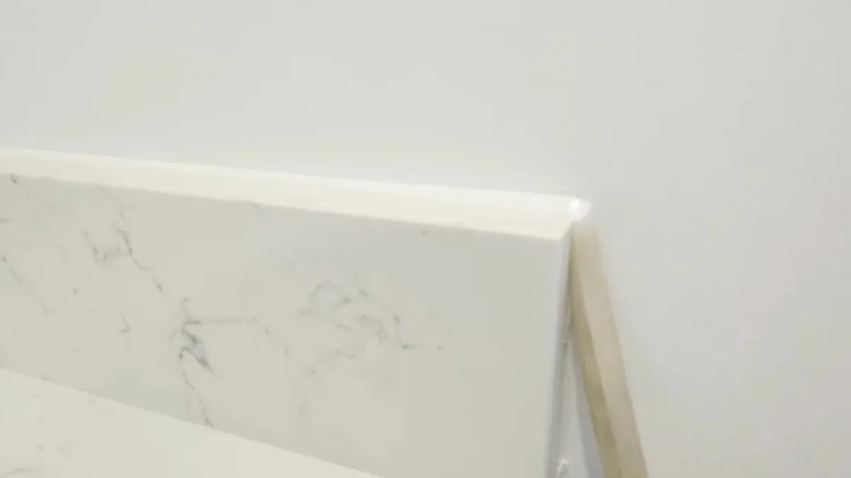Wooden tooling stick finishing a fresh white sealant bead at the end of a white quartz upstand where it meets a painted wall