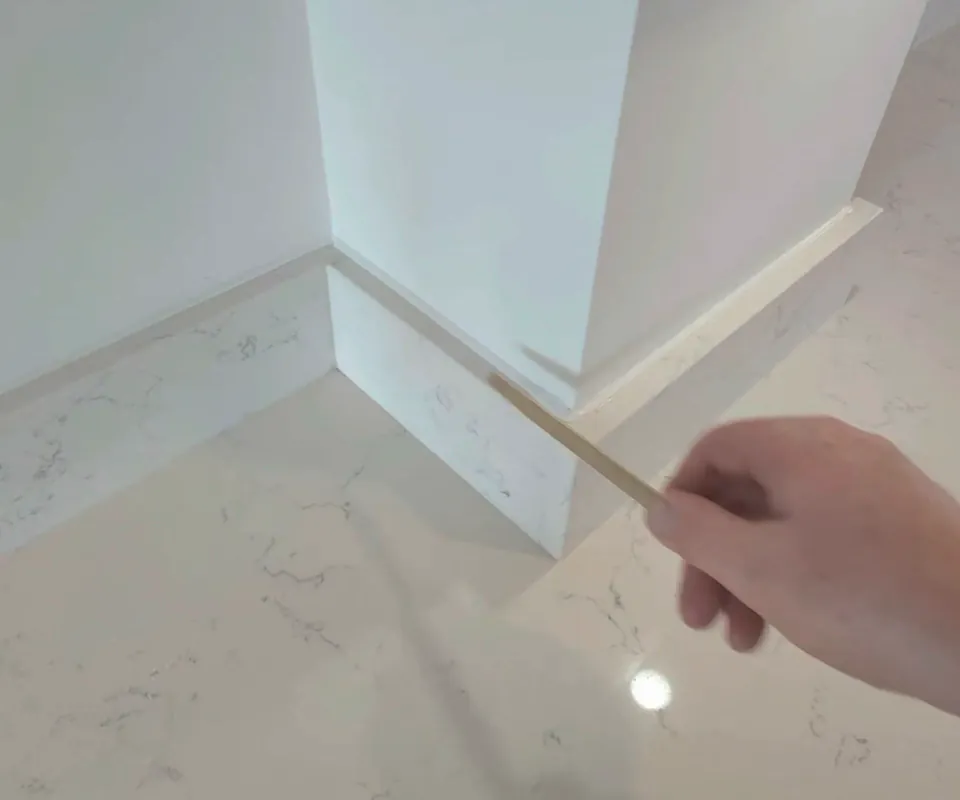 Wooden tooling stick smoothing sealant along the top of a white quartz upstand where it turns an external corner against a white wall