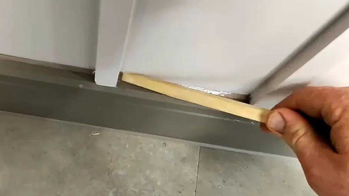 Applicator tooling a fresh sealant joint with a wooden spatula where a white door frame meets grey skirting above a stone-effect tiled floor